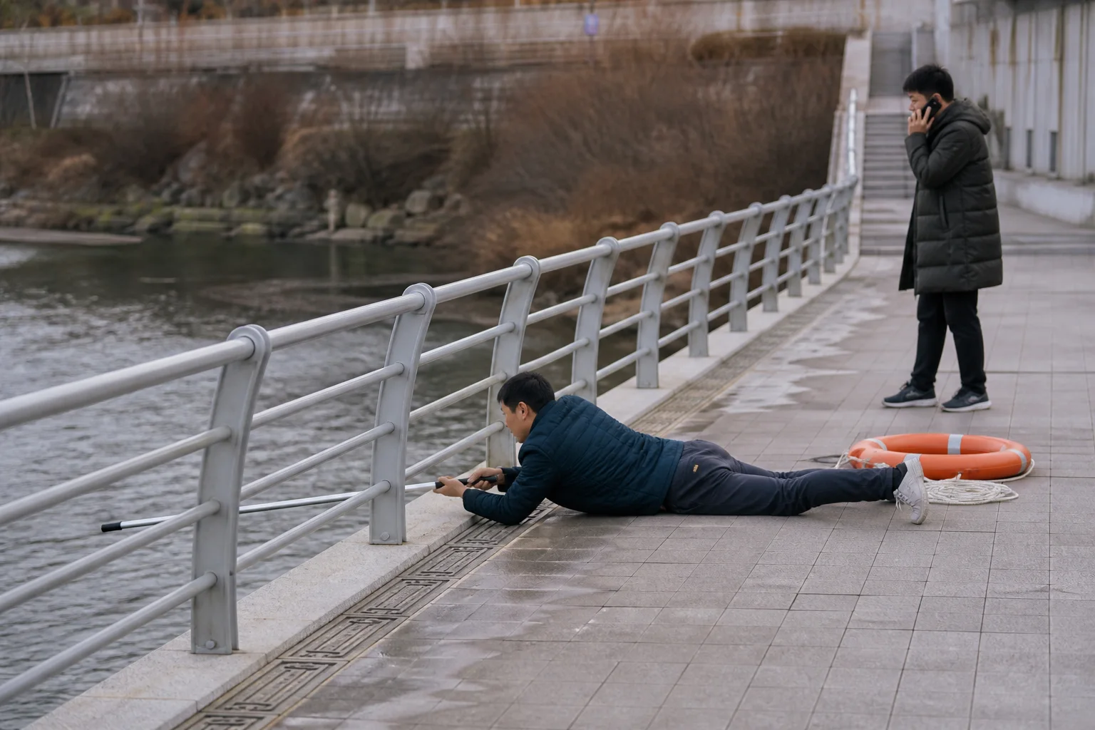 하천 난간 안쪽에서 한 사람이 바닥에 낮게 엎드려 긴 구조봉을 물 쪽으로 내밀고 다른 사람은 떨어져 신고하며 구명환을 준비한 상황 재구성 (AI 생성 삽화)