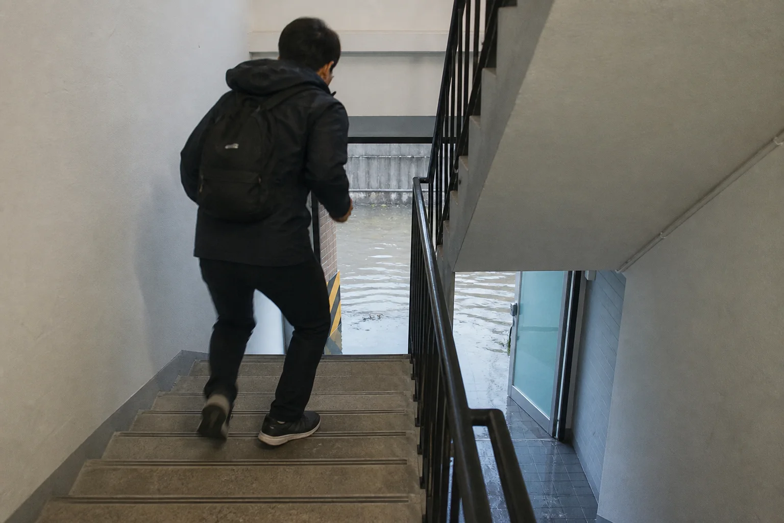 작은 배낭을 멘 주민이 마른 실내 계단으로 위층을 향해 오르고 물은 열린 1층 출입구 아래에만 머무는 상황 재구성 (AI 생성 삽화)